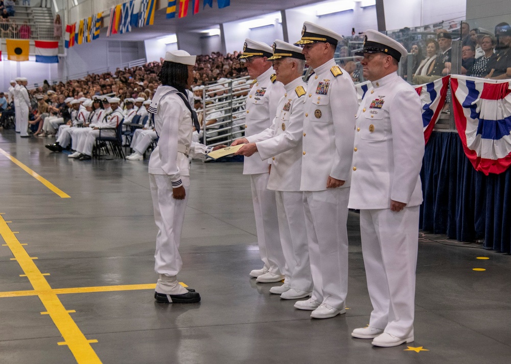 A U.S. Navy graduation or pass-in-review ceremony is taking place in a large indoor facility. A junior enlisted sailor in dress white uniform is receiving a document or certificate from a line of senior naval officers. A large audience of sailors and civilians observes from the bleachers in the background.