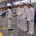 A U.S. Navy graduation or pass-in-review ceremony is taking place in a large indoor facility. A junior enlisted sailor in dress white uniform is receiving a document or certificate from a line of senior naval officers. A large audience of sailors and civilians observes from the bleachers in the background.