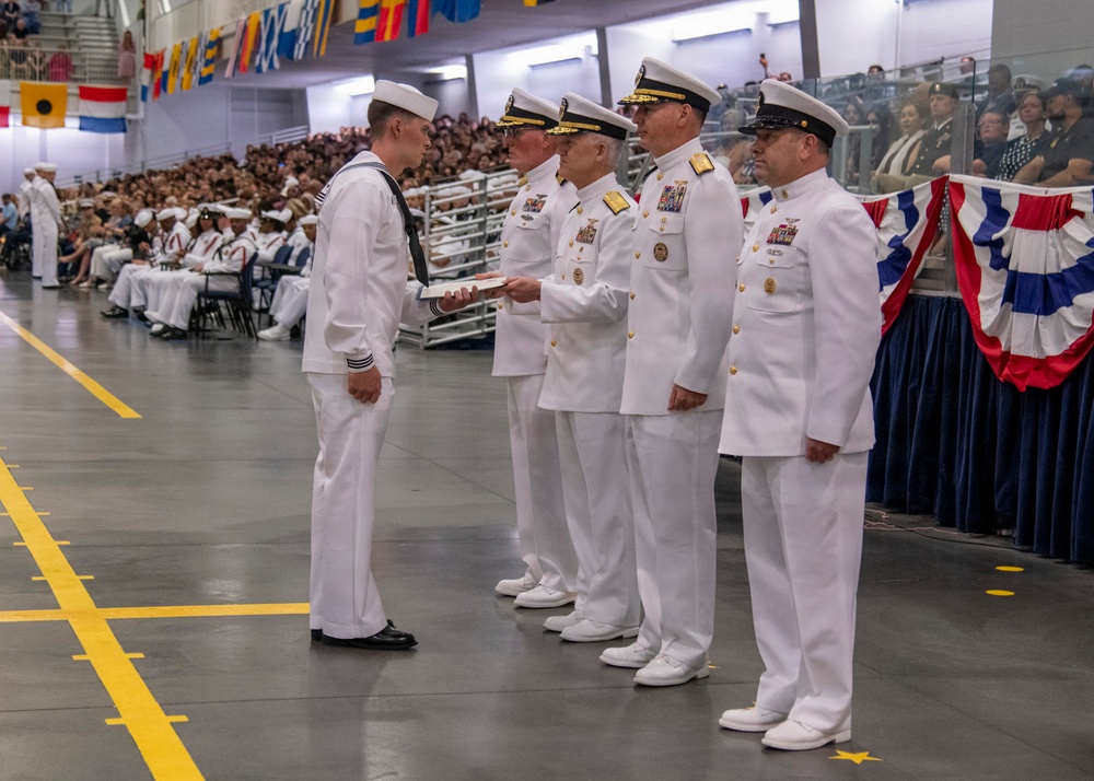 A U.S. Navy graduation or awards ceremony taking place in a large indoor facility, likely a recruit training command. A junior enlisted sailor in dress whites is receiving a document or certificate from a line of senior officers, also in dress white uniforms. A large audience of sailors and civilians is seated in bleachers in the background.