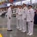 A U.S. Navy graduation or awards ceremony taking place in a large indoor facility, likely a recruit training command. A junior enlisted sailor in dress whites is receiving a document or certificate from a line of senior officers, also in dress white uniforms. A large audience of sailors and civilians is seated in bleachers in the background.