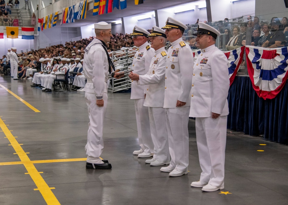 A U.S. Navy graduation or awards ceremony taking place in a large indoor facility, likely a recruit training command. A sailor in enlisted dress whites with a neckerchief stands before a line of four senior officers in officer dress whites, appearing to receive recognition or a handshake. The venue is filled with spectators in bleachers and seated rows, decorated with nautical signal flags and red, white, and blue bunting.