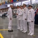 A U.S. Navy graduation or awards ceremony taking place in a large indoor facility, likely a recruit training command. A sailor in enlisted dress whites with a neckerchief stands before a line of four senior officers in officer dress whites, appearing to receive recognition or a handshake. The venue is filled with spectators in bleachers and seated rows, decorated with nautical signal flags and red, white, and blue bunting.