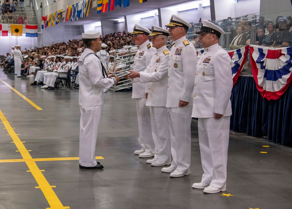 A U.S. Navy graduation or awards ceremony taking place in a large indoor facility, likely a recruit training command. A junior enlisted sailor in dress whites with a traditional sailor collar is receiving a plaque or award from a line of senior officers also dressed in white service uniforms. The venue is filled with seated recruits and civilian spectators in bleachers.