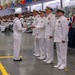 A U.S. Navy graduation or awards ceremony taking place in a large indoor facility, likely a recruit training command. A junior enlisted sailor in dress whites with a traditional sailor collar is receiving a plaque or award from a line of senior officers also dressed in white service uniforms. The venue is filled with seated recruits and civilian spectators in bleachers.