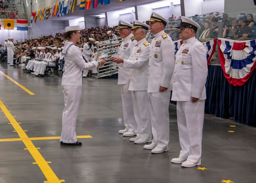 A formal U.S. Navy graduation or awards ceremony taking place in a large indoor facility, likely a drill hall or gymnasium. A junior enlisted sailor in dress whites is receiving something (likely a certificate or credential) from a line of senior officers, also in dress white uniforms. A large audience of sailors and civilians is seated in bleachers in the background.