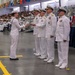 A formal U.S. Navy graduation or awards ceremony taking place in a large indoor facility, likely a drill hall or gymnasium. A junior enlisted sailor in dress whites is receiving something (likely a certificate or credential) from a line of senior officers, also in dress white uniforms. A large audience of sailors and civilians is seated in bleachers in the background.