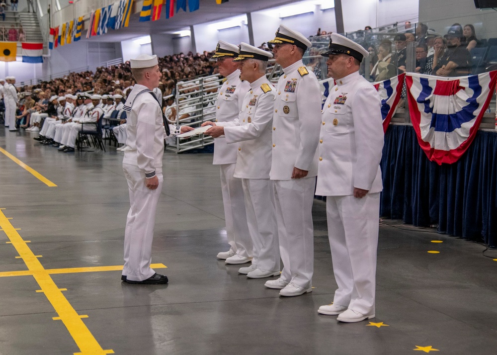 A U.S. Navy graduation or pass-in-review ceremony is taking place in a large indoor venue. A junior enlisted sailor in dress whites with a dixie cup hat is receiving a document or certificate from a line of senior officers in service dress white uniforms adorned with medals and gold insignia. A large audience of sailors and civilians observes from bleachers in the background.
