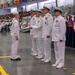 A U.S. Navy graduation or pass-in-review ceremony is taking place in a large indoor venue. A junior enlisted sailor in dress whites with a dixie cup hat is receiving a document or certificate from a line of senior officers in service dress white uniforms adorned with medals and gold insignia. A large audience of sailors and civilians observes from bleachers in the background.