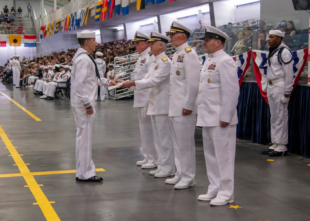 A U.S. Navy graduation or pass-in-review ceremony is taking place in a large indoor facility. A junior enlisted sailor in dress whites is receiving a document or certificate from a line of senior officers also in dress white uniforms. A large audience of sailors and civilians is visible in the background on bleachers.