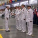 A U.S. Navy graduation or pass-in-review ceremony is taking place in a large indoor facility. A junior enlisted sailor in dress whites is receiving a document or certificate from a line of senior officers also in dress white uniforms. A large audience of sailors and civilians is visible in the background on bleachers.
