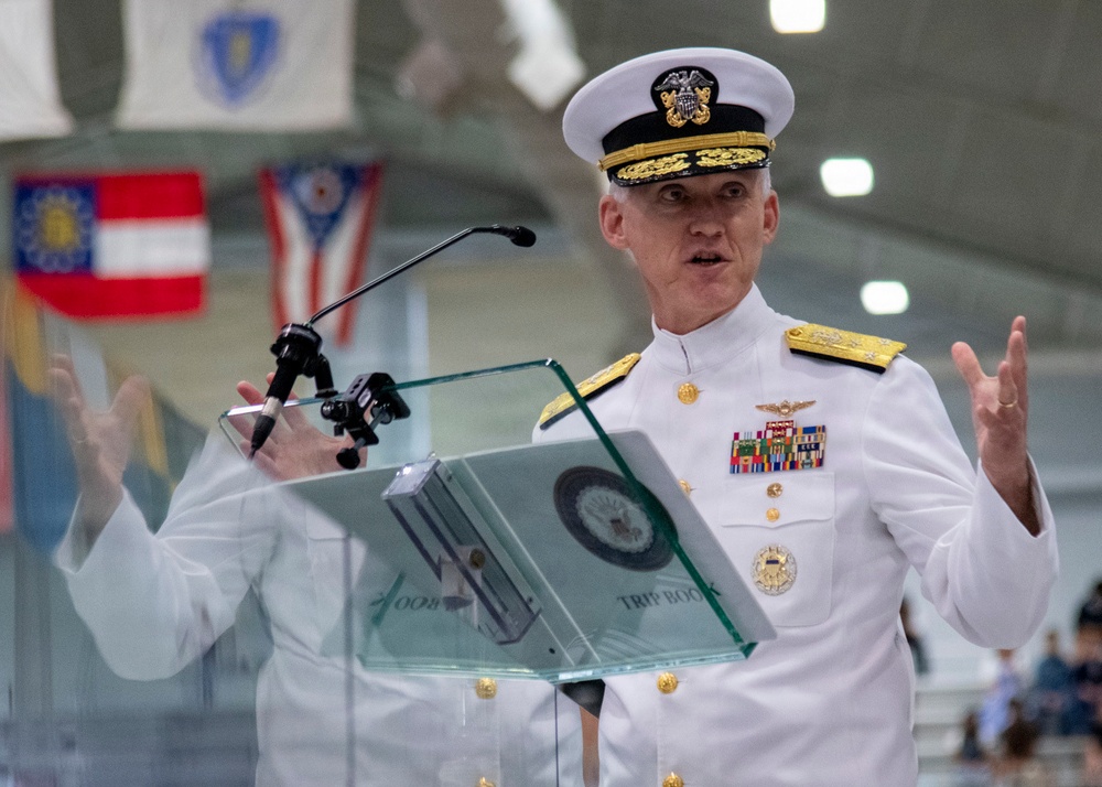 A senior U.S. Navy officer in dress white uniform is delivering a speech at a formal podium with a glass lectern. The officer displays multiple service ribbons, gold aviator wings, and shoulder boards indicating high flag officer rank. State flags are visible hanging in the background of what appears to be a large ceremonial hall.