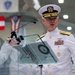 A senior U.S. Navy officer in dress white uniform is delivering a speech at a formal podium with a glass lectern. The officer displays multiple service ribbons, gold aviator wings, and shoulder boards indicating high flag officer rank. State flags are visible hanging in the background of what appears to be a large ceremonial hall.