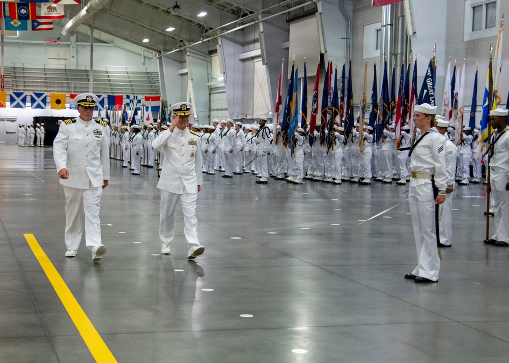 A formal U.S. Navy military ceremony is taking place inside a large indoor facility, likely a hangar or drill hall. Two senior naval officers in dress white uniforms are walking and rendering salutes while rows of sailors in white uniforms stand at attention. Multiple military color guard members holding state and unit flags are lined up in the background.