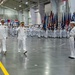 A formal U.S. Navy military ceremony is taking place inside a large indoor facility, likely a hangar or drill hall. Two senior naval officers in dress white uniforms are walking and rendering salutes while rows of sailors in white uniforms stand at attention. Multiple military color guard members holding state and unit flags are lined up in the background.