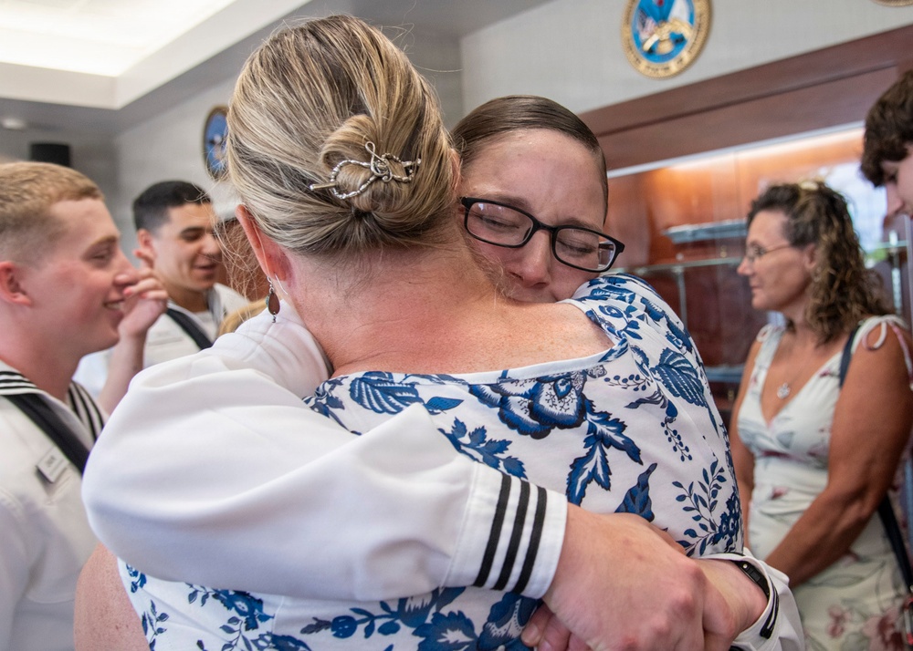 Two women are sharing an emotional embrace in what appears to be a military ceremony or official event. One woman is wearing a white uniform with black sleeve stripes, consistent with a naval officer's dress whites, while the other wears a blue and white floral dress. In the background, several other individuals including sailors in uniform are visible, along with an official seal mounted on the wall.