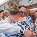 Two women are sharing an emotional embrace in what appears to be a military ceremony or official event. One woman is wearing a white uniform with black sleeve stripes, consistent with a naval officer's dress whites, while the other wears a blue and white floral dress. In the background, several other individuals including sailors in uniform are visible, along with an official seal mounted on the wall.