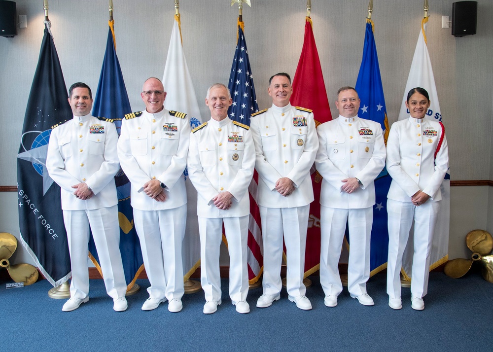 Six military personnel in white dress uniforms are posed for a formal group photograph in front of multiple service flags. The setting appears to be an official military ceremony or event, with flags including the U.S. Space Force, American flag, U.S. Army, and what appears to be a Coast Guard flag visible in the background. The individuals display various ranks indicated by insignia and ribbon/medal arrangements on their uniforms.