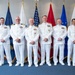 Six military personnel in white dress uniforms are posed for a formal group photograph in front of multiple service flags. The setting appears to be an official military ceremony or event, with flags including the U.S. Space Force, American flag, U.S. Army, and what appears to be a Coast Guard flag visible in the background. The individuals display various ranks indicated by insignia and ribbon/medal arrangements on their uniforms.