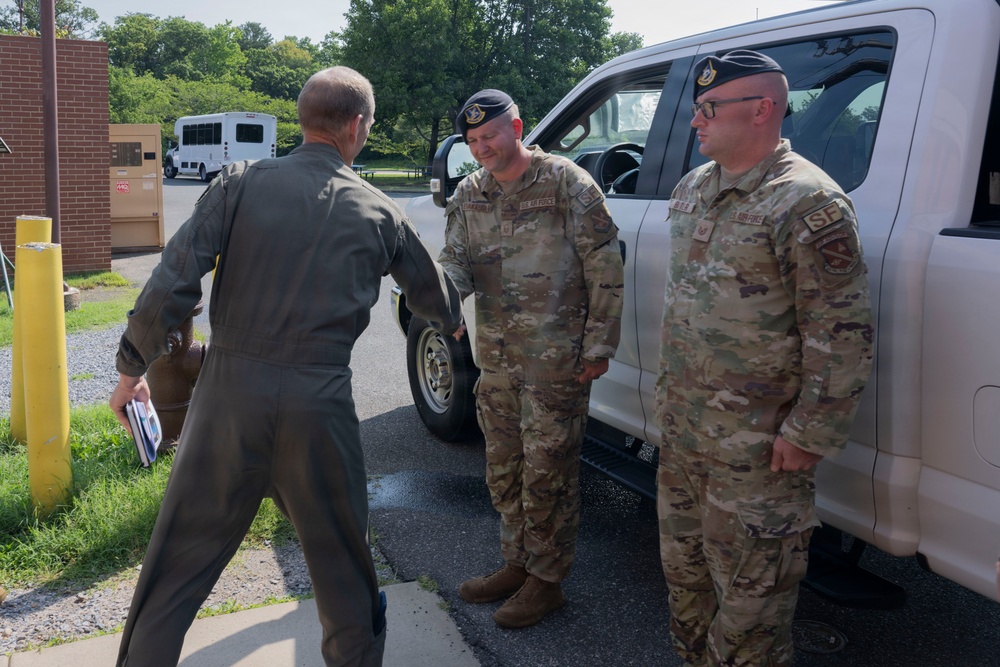 Three U.S. Air Force personnel are gathered outdoors near a white pickup truck on a military installation. One individual wearing an olive green flight suit is facing two others dressed in OCP (Operational Camouflage Pattern) uniforms with black berets. The interaction appears to be a casual conversation or briefing.