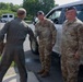 Three U.S. Air Force personnel are gathered outdoors near a white pickup truck on a military installation. One individual wearing an olive green flight suit is facing two others dressed in OCP (Operational Camouflage Pattern) uniforms with black berets. The interaction appears to be a casual conversation or briefing.