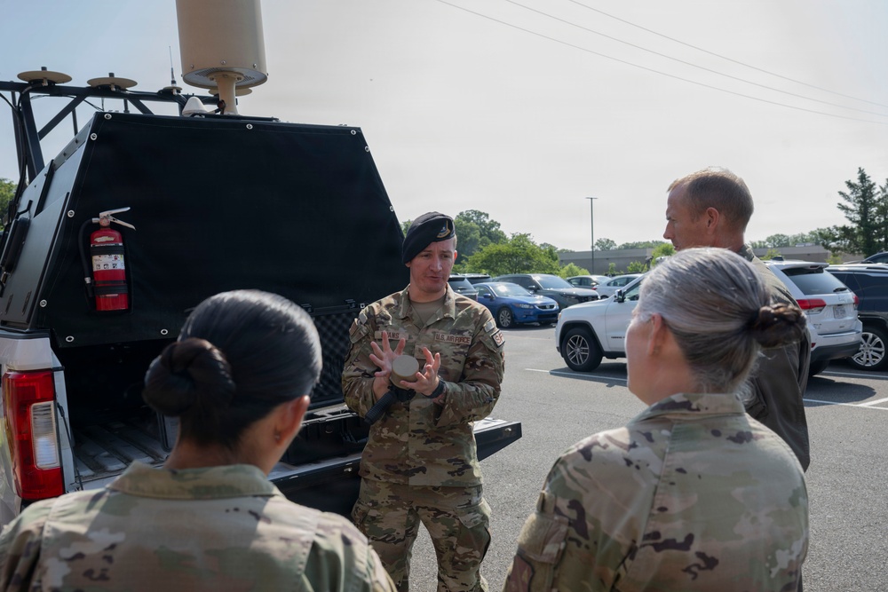 A U.S. Air Force personnel member wearing a black beret is conducting a briefing or demonstration for a small group of military personnel in camouflage uniforms in a parking lot. The instructor is holding a small circular object and appears to be explaining equipment mounted on the back of a tactical vehicle. The setting appears to be a military installation or facility during daytime.