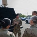 A U.S. Air Force personnel member wearing a black beret is conducting a briefing or demonstration for a small group of military personnel in camouflage uniforms in a parking lot. The instructor is holding a small circular object and appears to be explaining equipment mounted on the back of a tactical vehicle. The setting appears to be a military installation or facility during daytime.