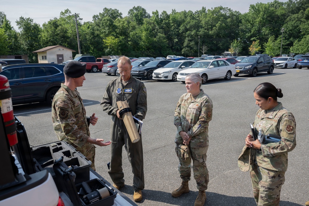 Four military personnel in uniform are gathered in a parking lot, appearing to conduct a briefing or equipment demonstration near the open trunk of a vehicle. One individual wearing a black beret and OCP camouflage uniform is gesturing and speaking, while the others listen attentively. One person in a flight suit is holding a cylindrical object, possibly a munition training aid or equipment component.