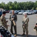 Four military personnel in uniform are gathered in a parking lot, appearing to conduct a briefing or equipment demonstration near the open trunk of a vehicle. One individual wearing a black beret and OCP camouflage uniform is gesturing and speaking, while the others listen attentively. One person in a flight suit is holding a cylindrical object, possibly a munition training aid or equipment component.