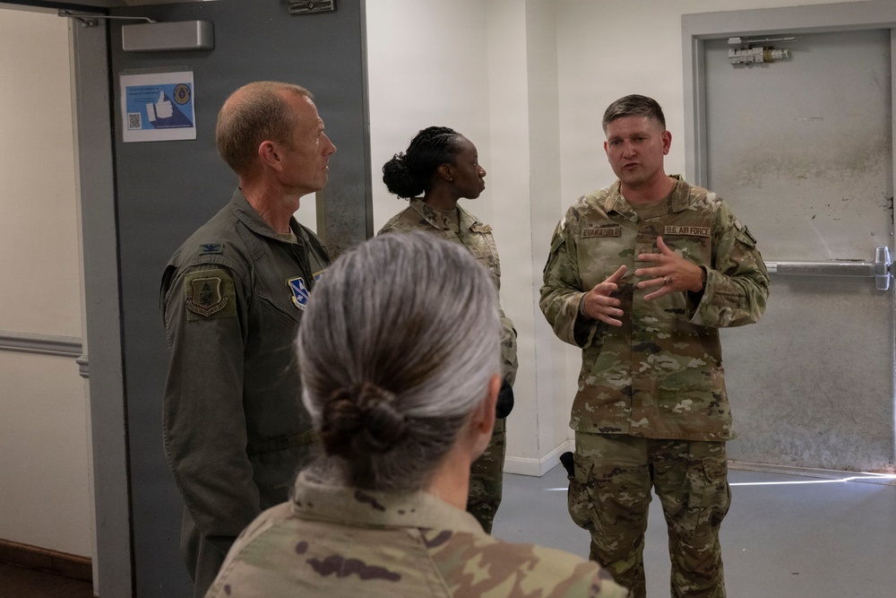 Four U.S. Air Force personnel are gathered in what appears to be an indoor military facility. A service member in OCP (Operational Camouflage Pattern) uniform is speaking and gesturing with their hands to the group. One individual is wearing an olive green flight suit with unit patches visible.