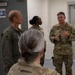 Four U.S. Air Force personnel are gathered in what appears to be an indoor military facility. A service member in OCP (Operational Camouflage Pattern) uniform is speaking and gesturing with their hands to the group. One individual is wearing an olive green flight suit with unit patches visible.