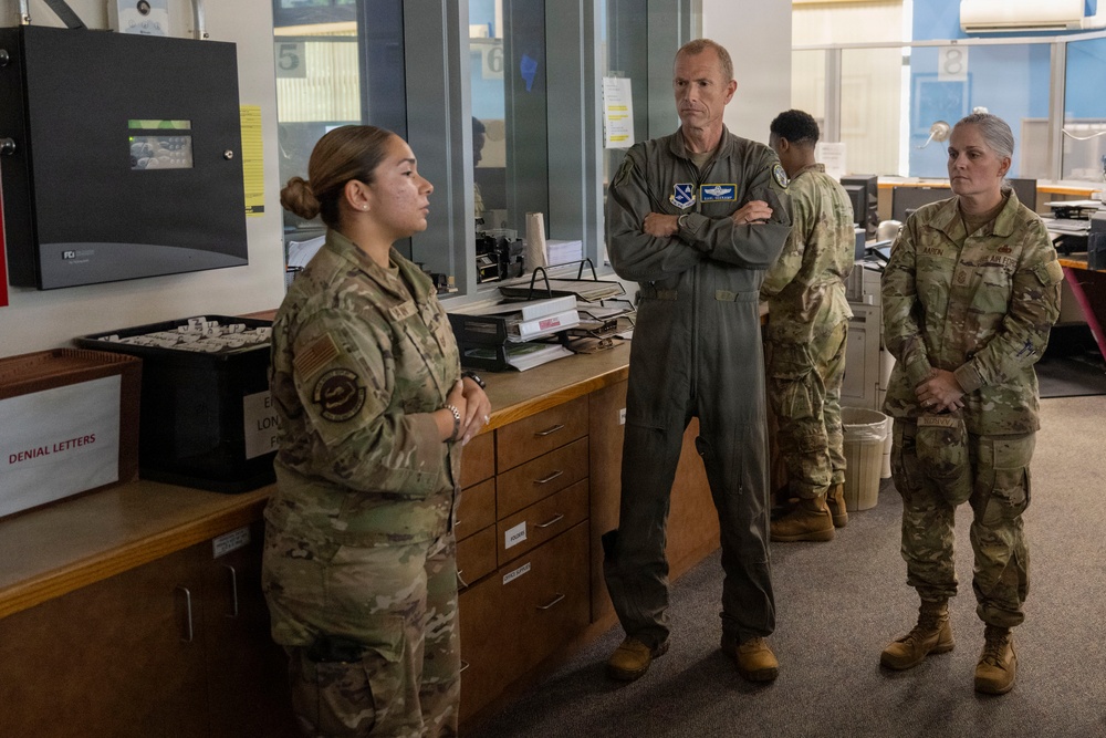 Four military personnel are gathered in what appears to be an administrative or operations office setting. A senior officer in an olive green flight suit stands with arms crossed, appearing to listen to a briefing from a junior enlisted member in camouflage uniform. A fourth service member is visible in the background at a workstation.