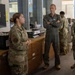Four military personnel are gathered in what appears to be an administrative or operations office setting. A senior officer in an olive green flight suit stands with arms crossed, appearing to listen to a briefing from a junior enlisted member in camouflage uniform. A fourth service member is visible in the background at a workstation.