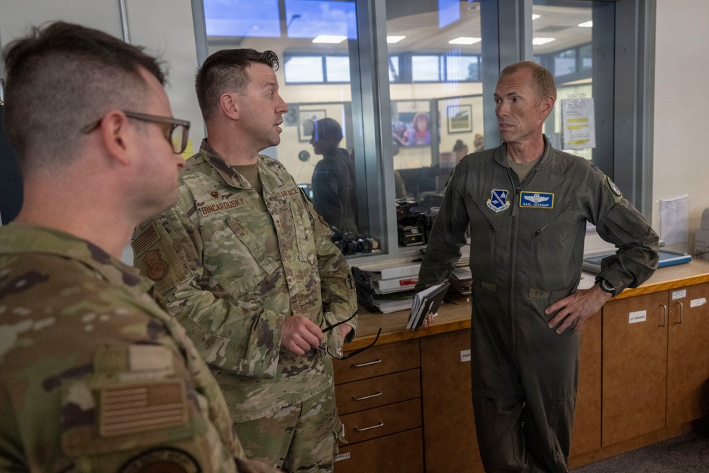 Three military personnel are engaged in a conversation inside what appears to be a military administrative office or operations center. Two individuals are wearing Army Combat Uniforms (ACU/OCP pattern), while the third is wearing an olive-green flight suit with unit patches and a name tag reading 'KARL DECKARD'. The setting suggests a briefing or informal discussion in a working military environment.