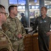 Three military personnel are engaged in a conversation inside what appears to be a military administrative office or operations center. Two individuals are wearing Army Combat Uniforms (ACU/OCP pattern), while the third is wearing an olive-green flight suit with unit patches and a name tag reading 'KARL DECKARD'. The setting suggests a briefing or informal discussion in a working military environment.