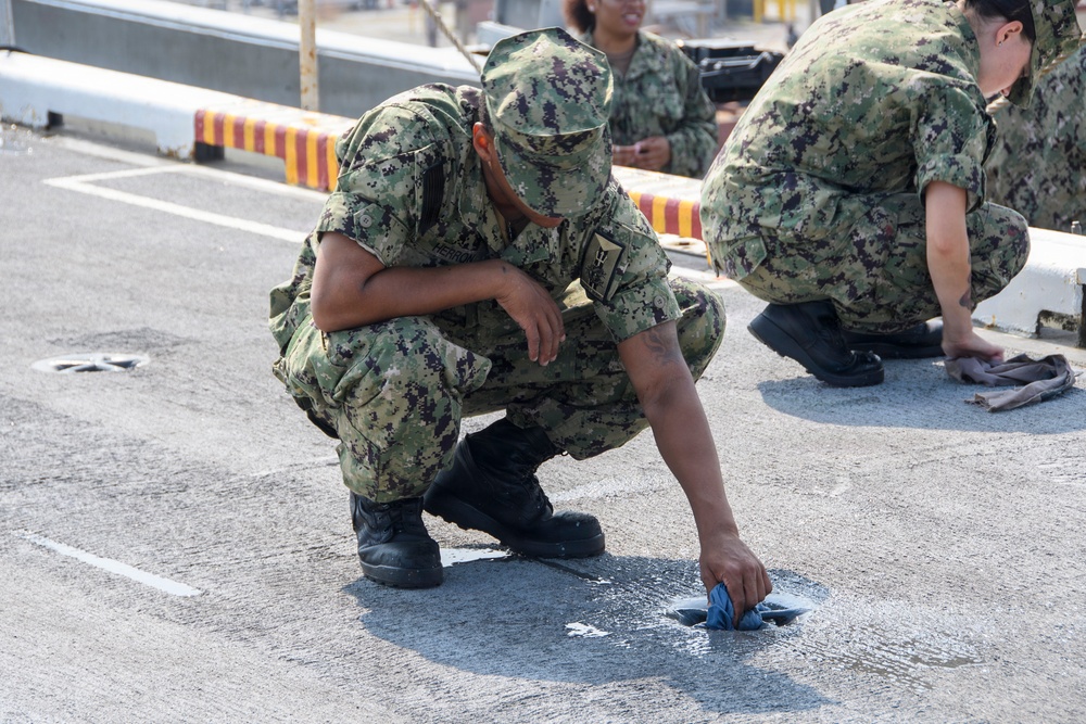 Multiple military personnel in digital camouflage uniforms are crouched down on a paved surface, cleaning or scrubbing the ground with cloths. The foreground figure is using a blue cloth to clean a wet or stained area on the pavement. A barrier with red and yellow stripes is visible in the background.