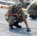Multiple military personnel in digital camouflage uniforms are crouched down on a paved surface, cleaning or scrubbing the ground with cloths. The foreground figure is using a blue cloth to clean a wet or stained area on the pavement. A barrier with red and yellow stripes is visible in the background.