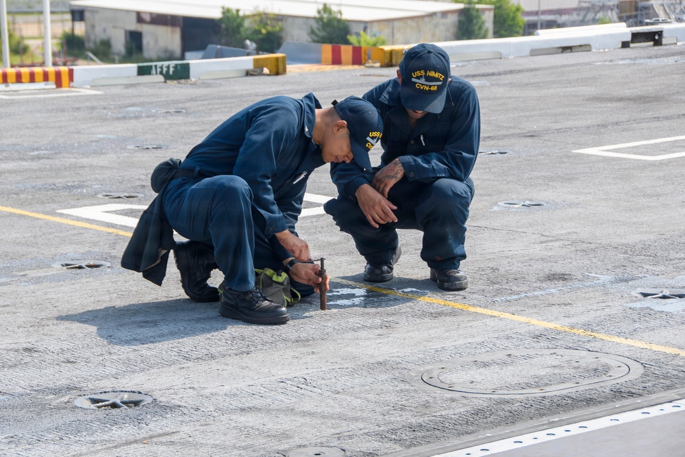 Two U.S. Navy sailors in dark blue coveralls are crouched on the flight deck of USS Nimitz (CVN-68), examining or working on a deck fitting or fastener. One sailor is kneeling and manipulating a tool near a deck component, while the other squats nearby observing. The setting is clearly a naval aircraft carrier flight deck, identifiable by the non-skid surface, tie-down rings, deck markings, and visible ship infrastructure in the background.
