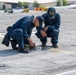 Two U.S. Navy sailors in dark blue coveralls are crouched on the flight deck of USS Nimitz (CVN-68), examining or working on a deck fitting or fastener. One sailor is kneeling and manipulating a tool near a deck component, while the other squats nearby observing. The setting is clearly a naval aircraft carrier flight deck, identifiable by the non-skid surface, tie-down rings, deck markings, and visible ship infrastructure in the background.