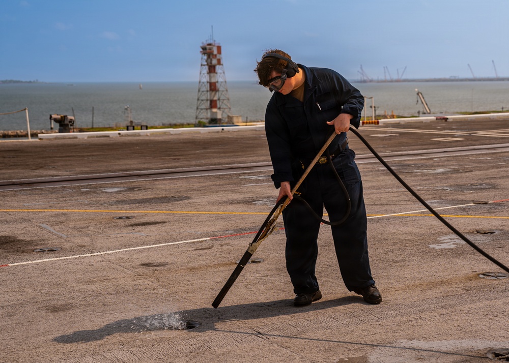 A person in dark navy coveralls and safety goggles is using a water hose with a long-handled nozzle to wash or clean a large flat surface, likely the flight deck of a naval vessel. The setting appears to be near a waterfront or port, with a red-and-white navigational tower and industrial cranes visible in the background. The work appears to be routine deck maintenance.