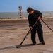 A person in dark navy coveralls and safety goggles is using a water hose with a long-handled nozzle to wash or clean a large flat surface, likely the flight deck of a naval vessel. The setting appears to be near a waterfront or port, with a red-and-white navigational tower and industrial cranes visible in the background. The work appears to be routine deck maintenance.