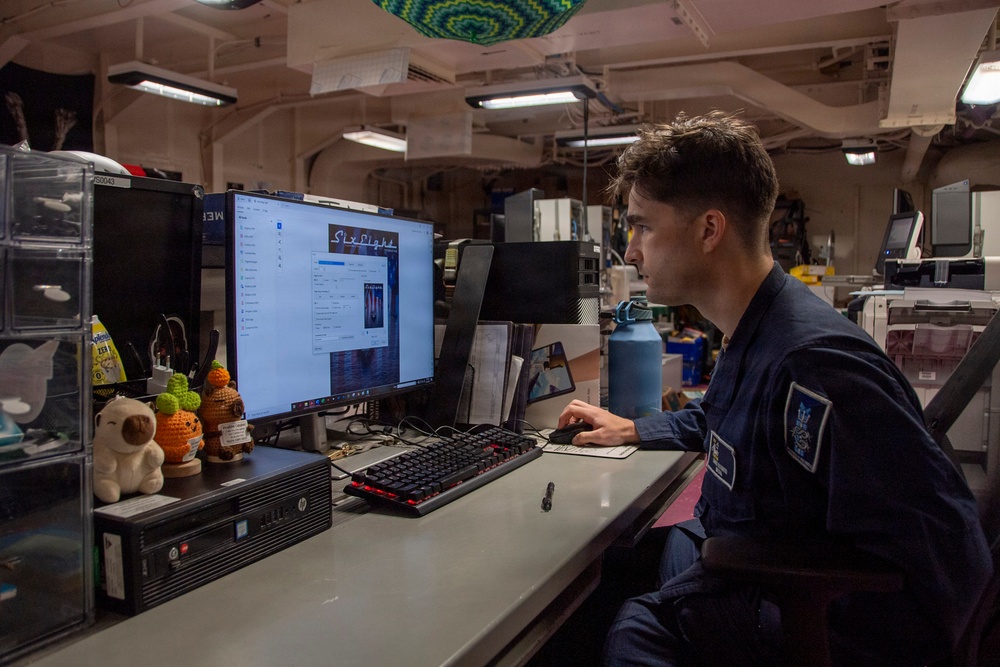 A young person in a dark blue military or naval uniform is seated at a workstation inside what appears to be a ship's interior compartment. They are working at an HP desktop computer displaying a Windows interface with what appears to be media or file management software. The workspace is personalized with small plush toys and figurines on the desk.