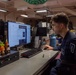 A young person in a dark blue military or naval uniform is seated at a workstation inside what appears to be a ship's interior compartment. They are working at an HP desktop computer displaying a Windows interface with what appears to be media or file management software. The workspace is personalized with small plush toys and figurines on the desk.