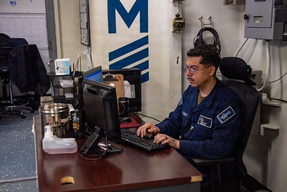 A U.S. Navy sailor in blue coveralls (NWU Type III) is seated at a desk working on a desktop computer in what appears to be an administrative office aboard a naval vessel. The workspace is cluttered with office supplies, beverages, and paperwork. A large blue 'M' insignia is visible on the wall behind the sailor.