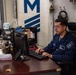 A U.S. Navy sailor in blue coveralls (NWU Type III) is seated at a desk working on a desktop computer in what appears to be an administrative office aboard a naval vessel. The workspace is cluttered with office supplies, beverages, and paperwork. A large blue 'M' insignia is visible on the wall behind the sailor.