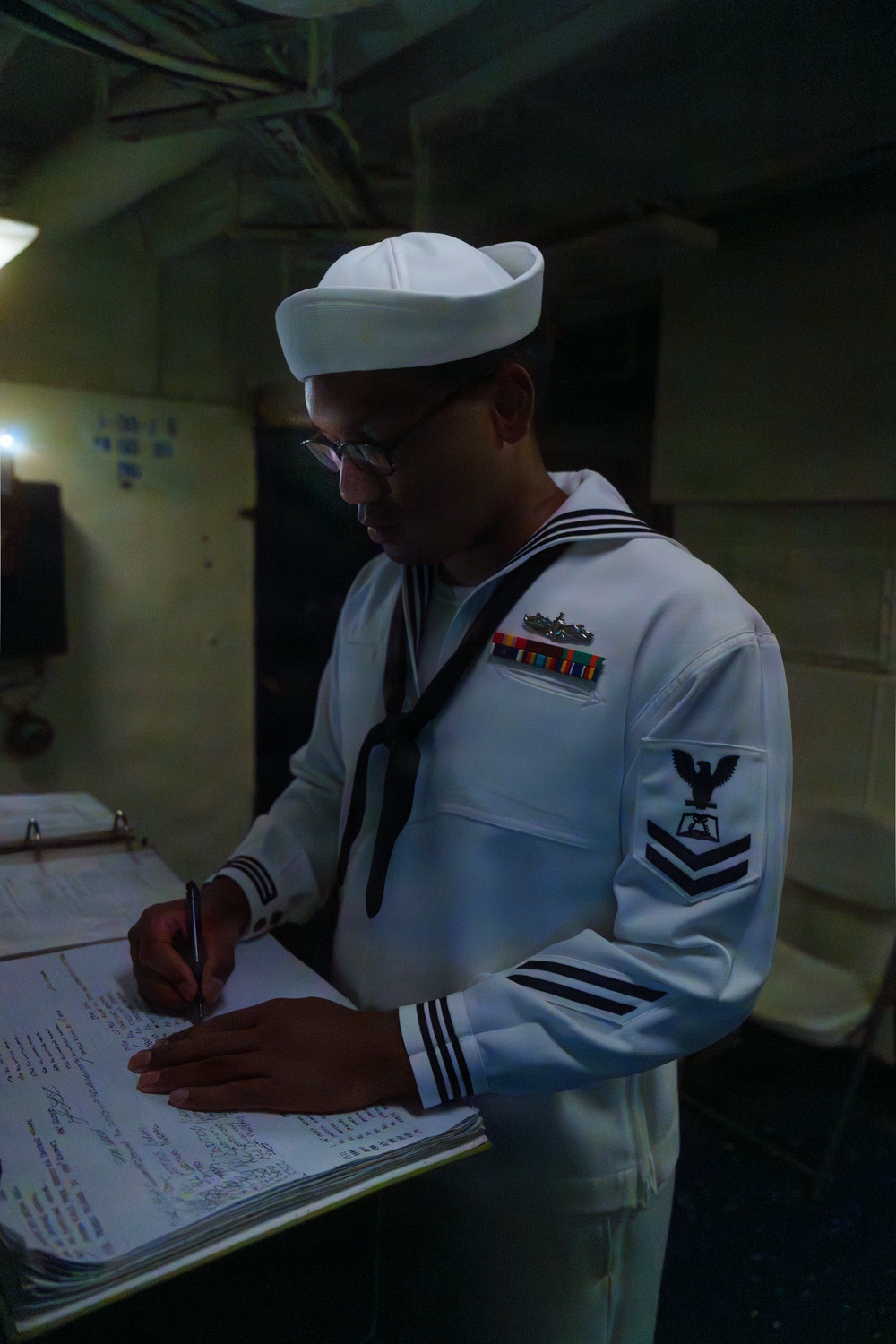 A U.S. Navy enlisted sailor in dress white uniform is standing at a counter writing in or signing a logbook or document. The sailor wears a Dixie cup white hat, black neckerchief, service ribbons, a warfare device pin, and a Petty Officer First Class rating badge on the left sleeve. The setting appears to be an interior compartment aboard a naval vessel.