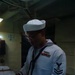 A U.S. Navy enlisted sailor in dress white uniform is standing at a counter writing in or signing a logbook or document. The sailor wears a Dixie cup white hat, black neckerchief, service ribbons, a warfare device pin, and a Petty Officer First Class rating badge on the left sleeve. The setting appears to be an interior compartment aboard a naval vessel.