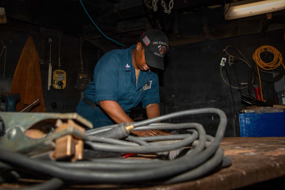 A Navy sailor wearing blue coveralls and a USS Nimitz 'Flying Eagles' cap is working at a workbench in what appears to be a ship's maintenance or engineering space. The sailor is handling heavy cables or hoses on a worn wooden workbench surrounded by tools and equipment. The setting is a dimly lit industrial workspace consistent with a naval vessel's interior.