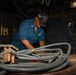 A Navy sailor wearing blue coveralls and a USS Nimitz 'Flying Eagles' cap is working at a workbench in what appears to be a ship's maintenance or engineering space. The sailor is handling heavy cables or hoses on a worn wooden workbench surrounded by tools and equipment. The setting is a dimly lit industrial workspace consistent with a naval vessel's interior.