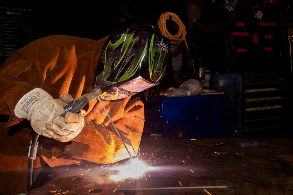 A welder is actively welding metal on a flat surface in what appears to be a workshop or fabrication shop. The person is wearing full protective gear including a Lincoln Electric auto-darkening welding helmet and leather welding gloves branded RIDGID. Bright sparks and arc light are visible at the weld point, illuminating the immediate work area.