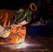A welder is actively welding metal on a flat surface in what appears to be a workshop or fabrication shop. The person is wearing full protective gear including a Lincoln Electric auto-darkening welding helmet and leather welding gloves branded RIDGID. Bright sparks and arc light are visible at the weld point, illuminating the immediate work area.